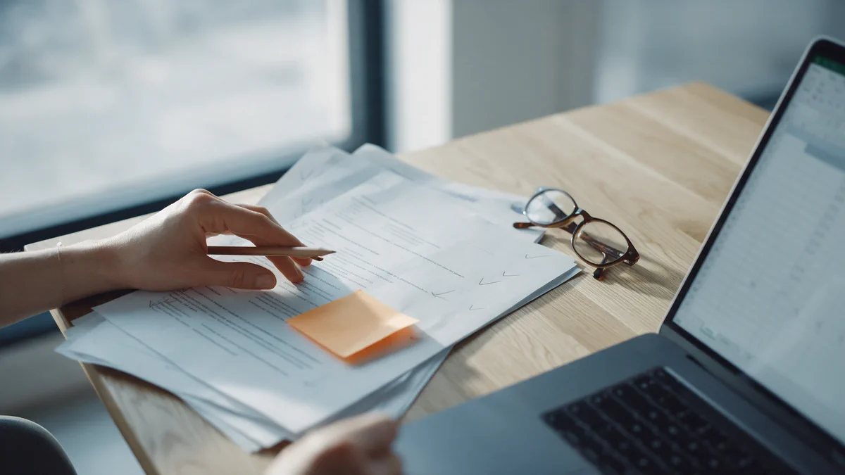 A desk with a stack of printed documents marked up in pencil, reading glasses and a laptop.